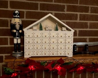 Op de afbeelding: Een witte houten adventskalender in de vorm van een huis met 24 genummerde lades. De kalender is versierd met kleine houten huisjes en bomen. Hij staat op een bakstenen schoorsteenmantel met een notenkrakerfiguur en een reeks rode lampjes.