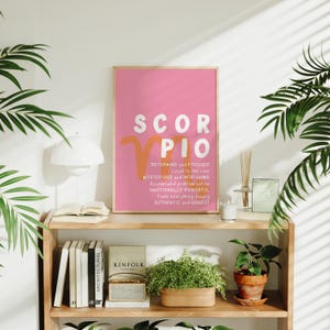 May include: A pink and orange poster with the zodiac sign Scorpio and a description of the Scorpio personality traits. The poster is framed and hanging on a wall above a wooden shelf with books and plants.