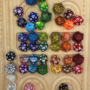 May include: A collection of multi-sided polyhedral dice in various colours and finishes, including translucent, solid, and glitter styles, arranged on a light wood board with etched measurement markings along the edges in centimetres.