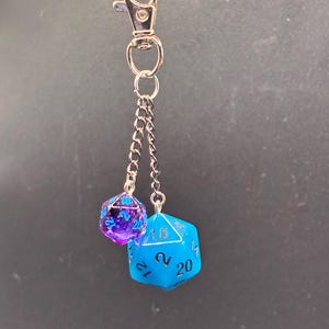 Puede incluir: Un llavero con un mosquetón de metal plateado y dos cadenas cortas. De las cadenas cuelgan dos dados poliédricos: uno más pequeño de color morado translúcido con números azules y otro más grande de color azul brillante opaco con números negros.
