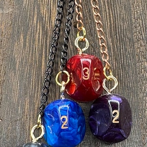 May include: Close-up of four colorful dice pendants hanging from chains. The dice are black, blue, red, and purple, each with a gold number. The chains are black and copper-colored.