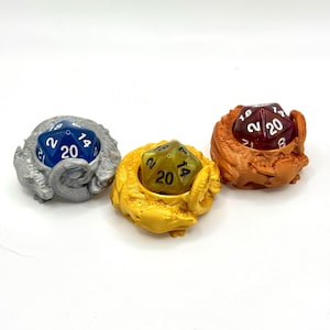 May include: Three dice holders, each with a D20 die inside. The holders are silver, gold, and orange, and are shaped like a ram's head. The dice are blue, yellow-green, and red-purple. The holders are 3D-printed.