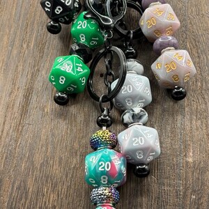 May include: A collection of four keychains featuring twenty-sided dice in various colors including black, green, purple, grey, and a teal-pink swirl. Each keychain incorporates black metal rings, small black beads, and iridescent textured spacer beads.