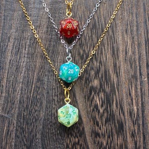 May include: Three layered necklaces featuring 20-sided die pendants. The top pendant is a glittery red die on a silver chain. The middle is a teal marbled die on a silver chain. The bottom is a pale green marbled die on a gold chain.