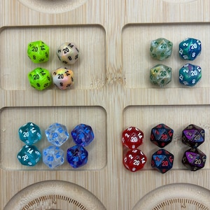 Puede incluir: Una colección de dados de veinte caras dispuestos en grupos sobre una bandeja de madera. El conjunto incluye dados de color verde neón, crema nacarado, verde jaspeado, azul, rojo y morado, con números blancos o rojos impresos en cada cara.