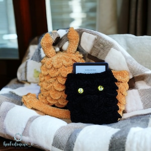 May include: A black crocheted e-reader cover with yellow eyes, a brown crocheted dragon, and a Kindle e-reader with the text "Kindle Unlimited" on the screen.