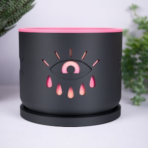 May include: Black planter with a pink rim and a cutout design of an eye with pink and orange accents.