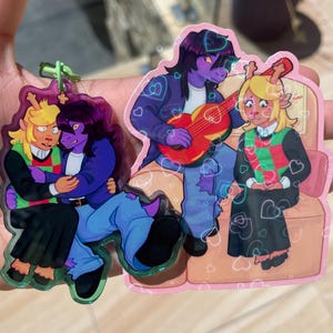 Może przedstawiać: Dwa przezroczyste breloki akrylowe z postaciami z kreskówek. Jeden brelok przedstawia dwie postacie obejmujące się, a drugi przedstawia trzy postacie, z których jedna gra na czerwonej gitarze. Postacie są w różnych pozach, z akcentami w kształcie serca.