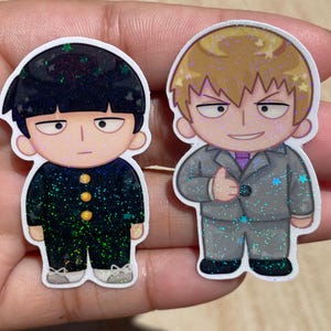 May include: Two glittery anime-style stickers. One shows a character in a black uniform with gold buttons, the other in a grey suit with a purple tie. Both have a cartoonish design with a white outline and star-shaped glitter.