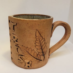 May include: A brown ceramic mug with a handle. The mug has a fern design and the word "TRISH" written on the side.