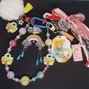 May include: A decorative keychain accessory featuring a beaded chain with iridescent and pastel spheres, a central rainbow charm with clouds, and various character-themed pendants. It includes a white pom-pom, a red and white gingham ribbon, and a pink tag.