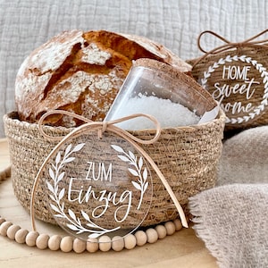Könnte beinhalten: Ein geflochtener Korb gefüllt mit einem Laib Brot und einem Glasgefäß mit weißem Zucker. Ein rundes, transparentes Acrylschild mit dem Text "zum Einzug" in weißer Schrift ist mit einem braunen Band am Korb befestigt. Das Schild ist von einem Kranz aus Blättern umgeben. Eine Holzperlenkette liegt im Vordergrund.