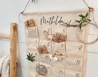 May include: A beige fabric advent calendar with pockets numbered 1-24. The name "Mathilda" is written across the top. Some pockets contain small wrapped gifts and wooden toys. The calendar is suspended from a wooden dowel.