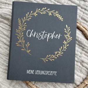 Könnte beinhalten: Ein graues, mit Leinen bezogenes Rezeptbuch mit einem goldenen Kranzdesign und dem Namen "Christopher" in weißer Schrift. Der Text "Meine Lieblingsrezepte" ist unten in weiß gedruckt.