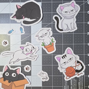 May include: A collection of die-cut vinyl stickers featuring various cartoon cats in different poses, including a black cat sleeping, a gray cat in a cone, a white cat in a plant pot, a white cat running, and a cat playing with a ball of yarn.