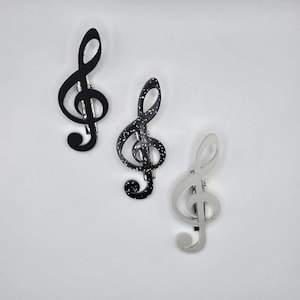 May include: Three treble clef hair clips in black, black with white speckles, and silver. The musical note-shaped clips are displayed against a white background. Each clip has a metal bar for securing the clip in the hair.
