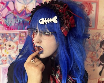 Può includere: Una persona con capelli blu vibranti indossa una fascia a quadri rossi e neri con bordo in pizzo e un piccolo fiocco. Una clip per capelli a forma di lisca di pesce bianca è fissata ai capelli. La persona indossa occhiali e un top nero.