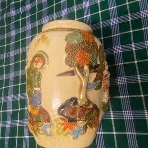 May include: A cream-coloured ceramic vase featuring a raised, multi-coloured relief design of a tree, foliage, and figures. The surface has a visible crackle glaze finish, with gold-toned accents on the base and rim, and intricate painted details.