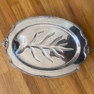 Puede incluir: Bandeja ovalada plateada con un diseño floral decorativo en el borde y un patrón tallado en forma de rama en el centro. La bandeja está sobre una superficie de madera.