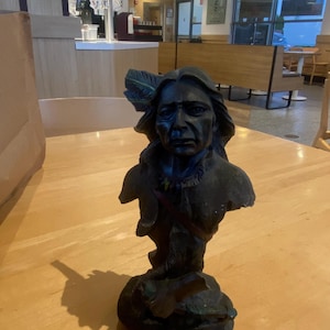 May include: A dark bronze-colored bust of a Native American man with long hair and a feather in his hair. The sculpture has a detailed face and is set on a textured base. The bust is displayed on a light wood table.