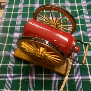 May include: A miniature red cannon model with wooden wheels and a wooden base. The wheels are yellow with black rims. A rope is attached to the cannon. The cannon is a decorative item.