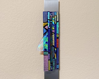 May include: A vertical wall-mounted mezuzah case featuring a rectangular brushed silver metal base. The front is decorated with a mosaic of fused glass strips in shades of blue, green, gold, and pink, with a single iridescent triangular glass accent.