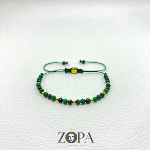 May include: A green beaded bracelet with gold accents. The bracelet features small, round, dark green beads interspersed with gold-colored beads. The adjustable cord is green. The brand name "ZOPA JEWELLERY" is printed below the bracelet.