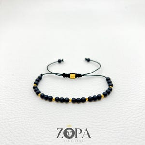 May include: Black beaded bracelet with gold-colored accents. The bracelet is made of small, round black beads interspersed with gold-colored beads. The bracelet is on a white surface. The brand name "ZOPA JEWELLERY" is visible at the bottom.