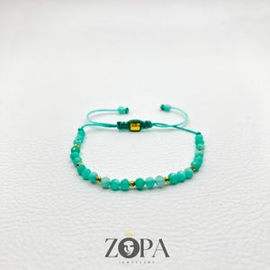 May include: A turquoise beaded bracelet with gold accents. The adjustable bracelet features small, round turquoise beads interspersed with gold-colored beads. The bracelet is on a white surface, with the brand logo "ZOPA JEWELRY" visible below.