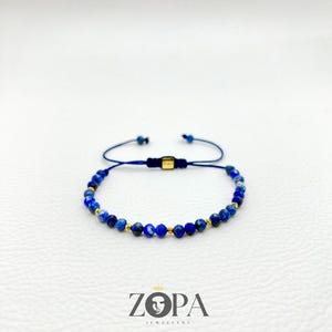 May include: A blue beaded bracelet with gold accents and a navy blue adjustable cord. The bracelet features small, round, dark blue beads. The brand name "ZOPA JEWELLERY" is printed in black below the bracelet.