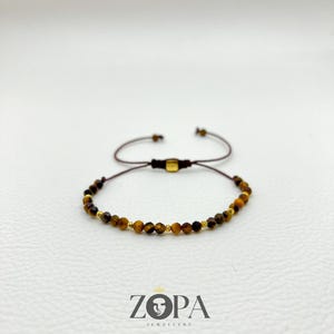 May include: A brown beaded bracelet with gold accents. The bracelet features small, faceted beads and is adjustable with a sliding knot closure. The brand name "ZOPA JEWELLERY" is displayed below the bracelet.