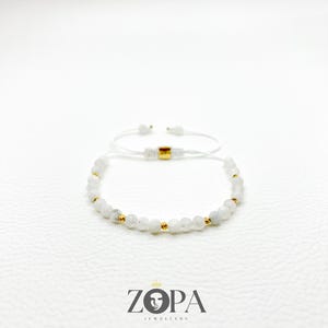 May include: White beaded bracelet with gold accents and a gold square bead. The bracelet is on a white background. The brand name "ZOPA JEWELLERY" is printed in black below the bracelet.