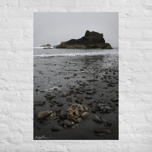 May include: A coastal landscape print featuring a rocky beach under a cloudy sky. The foreground shows a skull-like rock formation among pebbles. Waves gently lap the shore, with a large rock formation in the distance. The overall color palette is muted, with grays and browns.
