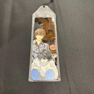 May include: A clear plastic bookmark featuring two anime characters. One character has light brown hair and is wearing a gray shirt. The other character has black hair and is wearing a white shirt and blue pants. A black tassel hangs from the top.