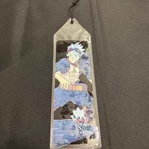May include: A clear plastic bookmark featuring a colorful anime character illustration. The character has spiky white hair, a headband, and is wearing a blue outfit. A black tassel hangs from the top of the bookmark.