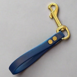 May include: A blue leather dog leash handle with a gold-colored metal clasp and rivets. The handle is a deep blue color and appears to be made of high-quality leather. The clasp is a bright gold color.