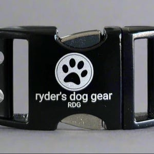 Op de afbeelding: Zwarte hondenriemgesp met een zilveren pootafdruklogo en de tekst "ryder's dog gear". De gesp heeft aan elke kant twee zilveren klinknagels en een metalen sluiting. Ontworpen voor dierenaccessoires.