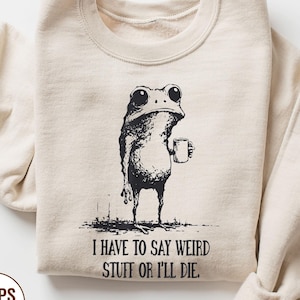May include: Sand-colored sweatshirt with a black graphic of a frog holding a mug. The text "I HAVE TO SAY WEIRD STUFF OR I'LL DIE" is below the frog. The sweatshirt is a neutral color and has long sleeves.