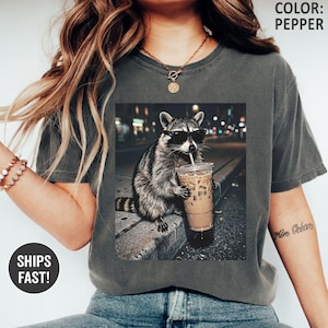 May include: Pepper-colored t-shirt featuring a graphic of a raccoon wearing sunglasses and drinking iced coffee. The raccoon is set against a blurred city night scene. The shirt has a relaxed fit.