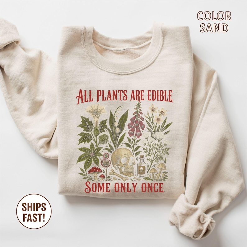 Pu&ograve; includere: Felpa color sabbia con un'illustrazione botanica e il testo rosso "ALL PLANTS ARE EDIBLE SOME ONLY ONCE". Il design include un teschio, varie piante e funghi.