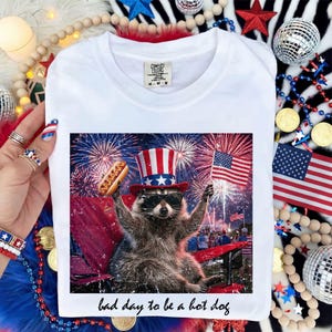 Puede incluir: Camiseta blanca con un dise&ntilde;o de un mapache con sombrero de T&iacute;o Sam y gafas de sol, sosteniendo un perrito caliente y una bandera estadounidense, con fuegos artificiales de fondo. El texto "bad day to be a hot dog" est&aacute; debajo.