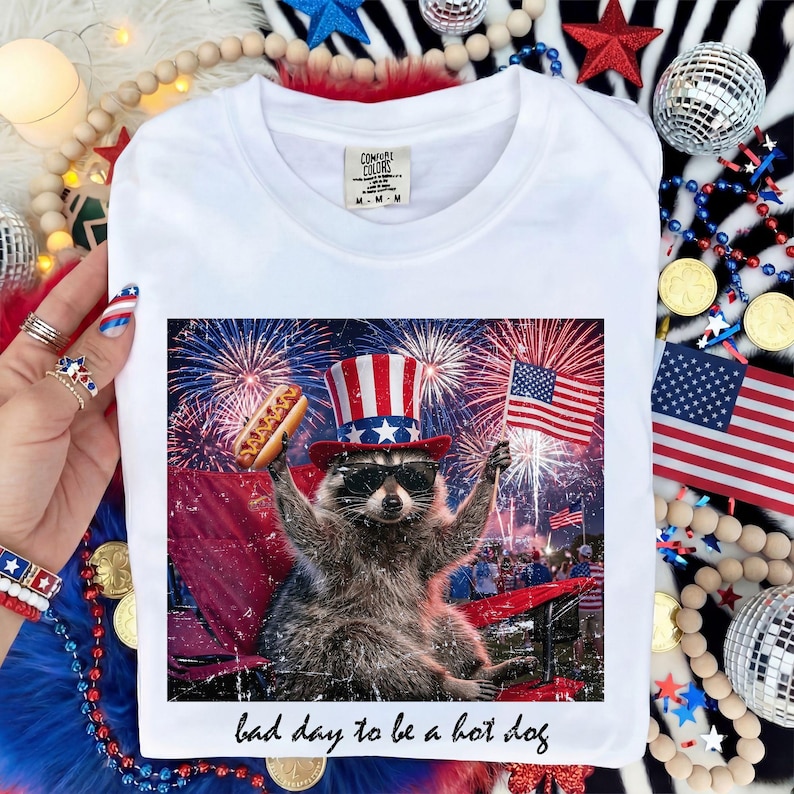 Puede incluir: Camiseta blanca con un dise&ntilde;o de un mapache con sombrero de T&iacute;o Sam y gafas de sol, sosteniendo un perrito caliente y una bandera estadounidense, con fuegos artificiales de fondo. El texto "bad day to be a hot dog" est&aacute; debajo.