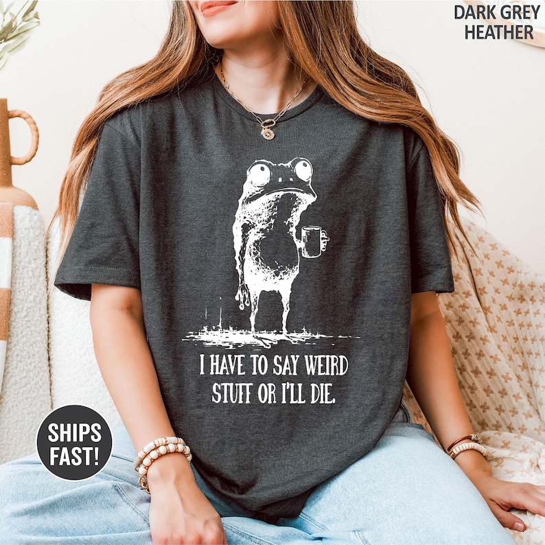 May include: Dark grey heather t-shirt featuring a white illustration of a frog holding a mug. The text "I HAVE TO SAY WEIRD STUFF OR I'LL DIE" is printed below the frog. The shirt is a casual wear item.
