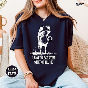 May include: Navy blue t-shirt featuring a white illustration of a frog holding a mug. The frog has a quirky expression. The text "I HAVE TO SAY WEIRD STUFF OR I'LL DIE" is printed below the frog.