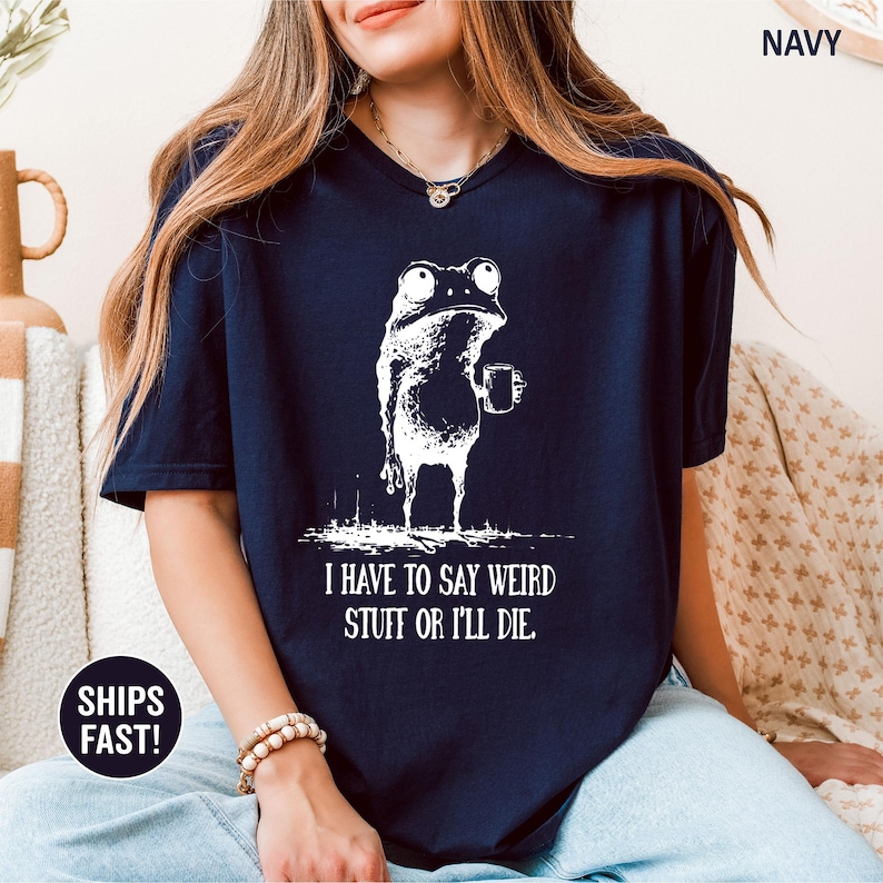 May include: Navy blue t-shirt featuring a white illustration of a frog holding a mug. The frog has a quirky expression. The text "I HAVE TO SAY WEIRD STUFF OR I'LL DIE" is printed below the frog.