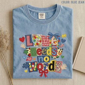 May include: Blue jean-colored t-shirt with the phrase "LOVE needs no WORD" in colorful, patchwork-style letters and shapes. The design includes hearts, puzzle pieces, and a bow. The shirt is folded on a neutral surface.
