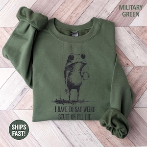 May include: Military green sweatshirt with a black graphic of a frog holding a mug. The text "I HAVE TO SAY WEIRD STUFF OR I'LL DIE" is printed below the frog. The sweatshirt has long sleeves and a crew neck.