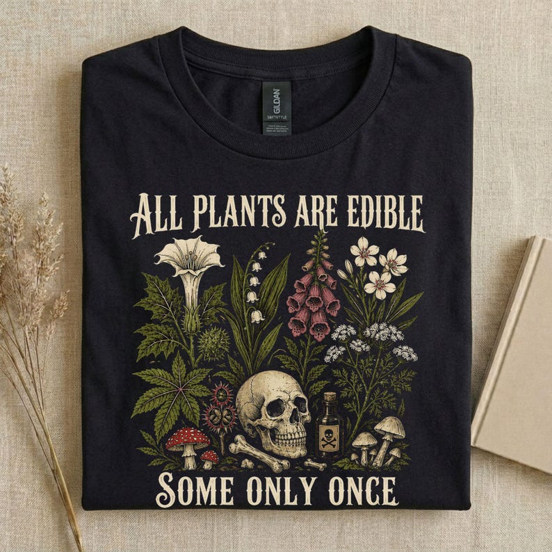 Pu&ograve; includere: T-shirt nero con un'illustrazione botanica raffigurante un teschio, varie piante e la scritta "ALL PLANTS ARE EDIBLE SOME ONLY ONCE". Il design include funghi, fiori e una bottiglia di veleno.