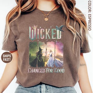 May include: Espresso-colored t-shirt featuring the word "Wicked" in a colorful, distressed font. Below, the phrase "Changed for Good" is displayed. The shirt showcases a graphic of two characters from the musical, one in green and the other in a blue gown.