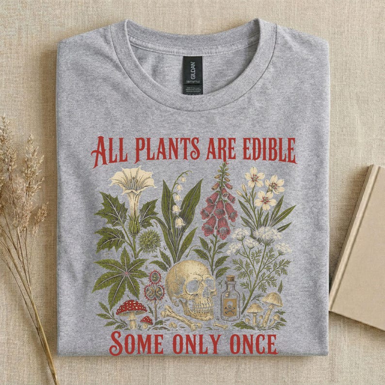 Pu&ograve; includere: Una maglietta grigia con un'illustrazione botanica e la scritta rossa "ALL PLANTS ARE EDIBLE SOME ONLY ONCE". Il design include varie piante, un teschio e una piccola bottiglia.